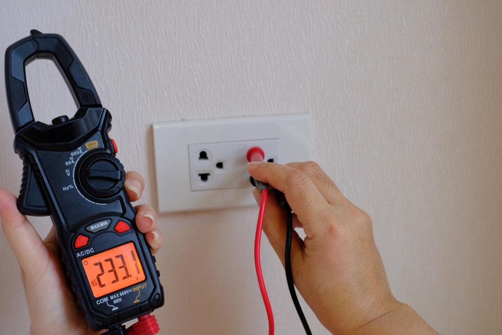 An electrician is a hand holding on the digital multimeter into a hole of an electric socket
